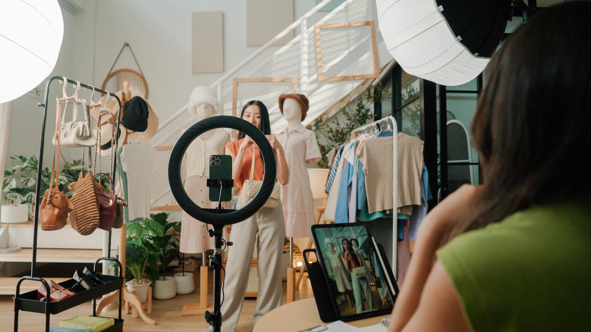 Young Asian woman overlooking a live selling session of  clothing store and assisting her friend in front of the camera.